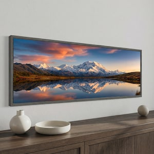 May include: A framed landscape print showing a mountain range reflected in a lake. The sky has orange, pink, and blue hues. The frame is dark grey. A white ceramic vase and bowl are on a wooden surface. The print is a scenic view.