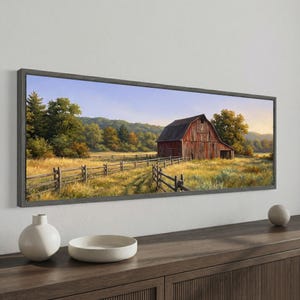 May include: A framed landscape painting depicts a rustic red barn in a field, with a wooden fence and trees. The artwork has a gray frame and is displayed on a wooden cabinet. The scene evokes a sense of rural tranquility.