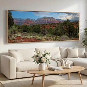 May include: A panoramic landscape wall art print featuring red rock mountains under a blue sky with clouds, surrounded by green desert vegetation, shrubs, and small white flowers, framed in a light wood border and displayed above a sofa.