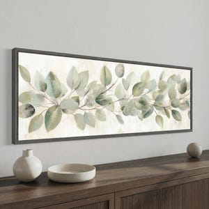May include: A framed botanical print featuring a watercolor depiction of eucalyptus leaves in shades of green and gray. The artwork is set against a cream-colored background and framed in a dark gray border. The print is displayed above a wooden cabinet.