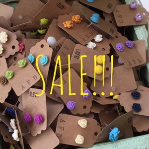 May include: A collection of colorful floral stud earrings on brown tags, displayed in a green container. The word "SALE!!!" is written in yellow across the image. The earrings feature various colors and styles.