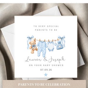 May include: A square baby shower greeting card featuring a watercolour illustration of a teddy bear, a blue patterned onesie, a striped bodysuit, a heart, and a blue knit hat hanging on a clothesline with blue bows. Text reads: TO VERY SPECIAL PARENTS TO BE, Lauren & Joseph, ON YOUR BABY SHOWER, 07.09.26, and a blue star.