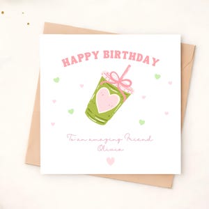 May include: A square white greeting card featuring the pink text HAPPY BIRTHDAY at the top and a green iced drink illustration with a pink heart and straw in the centre. Below, the text reads To an amazing Friend Olivia with small hearts.