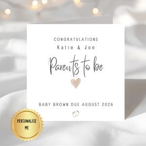 Può includere: Una carta bianca con il testo "Congratulations Katie & Joe Parents to be" e "Baby Brown Due August 2026". Un sigillo dorato con "Personalise Me" è nell'angolo in basso a sinistra. Un cuore dorato e piedini di bambino sono anche sulla carta.