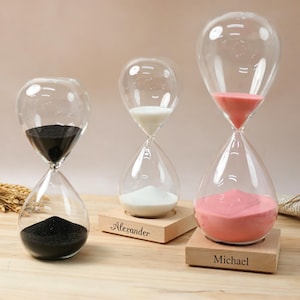 May include: Three clear glass hourglasses with sand in black, white, and pink. Two hourglasses feature light-coloured wooden bases with the engraved names Alexander and Michael. The hourglasses are arranged in a row on a flat surface.