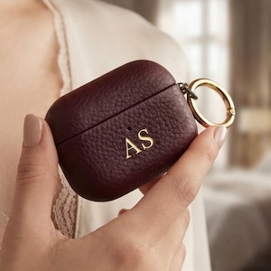 May include: A dark burgundy, pebbled-texture protective case for wireless earbuds. The case features a gold-toned metal ring attachment on the side and the gold-embossed initials AS centered on the front surface of the lid and base.