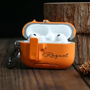 Può includere: Custodia protettiva arancione per auricolari wireless con il nome "Raquel" in corsivo. La custodia è aperta, rivelando auricolari bianchi all'interno. Un portachiavi argentato è attaccato.