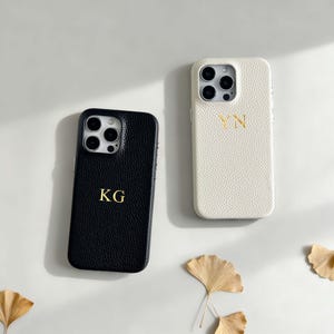 以下が含まれることがあります： 2つのパーソナライズされた携帯電話ケース。1つは金色のイニシャル「KG」が入った黒色、もう1つは金色のイニシャル「VN」が入った白色です。どちらのケースも革のような質感で、装飾的な葉のあるニュートラルな背景に配置されています。