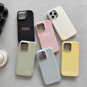 Puede incluir: Una colección de fundas personalizadas para teléfonos en varios colores: negro, rosa, blanco, verde, azul y amarillo. Cada funda presenta iniciales doradas. Las fundas tienen una apariencia texturizada similar al cuero.