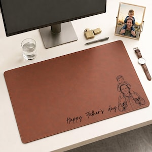 May include: A brown leather desk mat with a sketch of a father and child, and the text "Happy Father's Day." A framed photo and a watch are also on the desk.