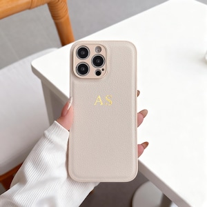 Könnte beinhalten: Hellbeige Handyhülle mit strukturierter Oberfläche. Das Gehäuse hat ein goldfarbenes Monogramm der Buchstaben "AS" und einen Kameraausschnitt. Die Handyhülle ist für ein Smartphone konzipiert.