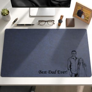 Op de afbeelding: Een marineblauwe bureaumat met een getextureerd oppervlak. De mat heeft een zwart-wit illustratie van een vader en kind, met de tekst "Best Dad Ever!". Een pen, bril, horloge en portemonnee liggen op het bureau.