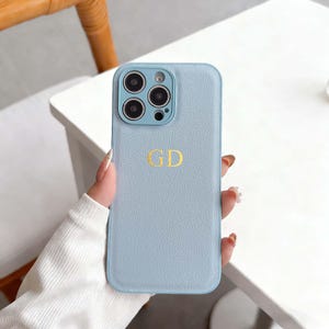 Op de afbeelding: Lichtblauwe telefoonhoes met de gouden initialen "GD". De hoes heeft een textuuroppervlak en is ontworpen voor een smartphone met meerdere cameralenzen.