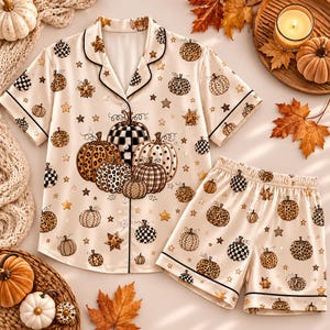 May include: A cream-colored pajama set featuring a short-sleeved button-down shirt and matching shorts. The fabric is decorated with a repeating pattern of pumpkins in various textures, including leopard print and checkered designs, interspersed with small stars.