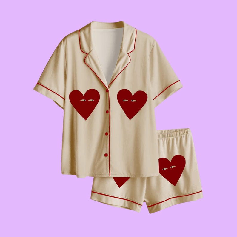 May include: Cream-colored pajama set with red trim and heart graphics. The short-sleeved top has two large heart designs with eyes, and the shorts feature a matching heart. The set is button-down and has a relaxed fit.