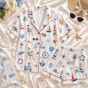 May include: A white short-sleeve pajama set with red piping, featuring a nautical print of lighthouses, sailboats, scooters, seashells, starfish, cherries, and people relaxing in chairs. The set includes a button-down shirt and matching shorts.