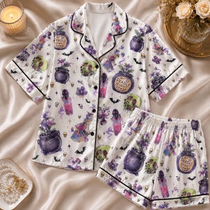 May include: A white short-sleeve button-down pajama shirt and matching shorts set featuring a repeating print of purple cauldrons, green poison apples, pink potion bottles, small skulls, and black bats. The text Deadly Night Shade is visible.