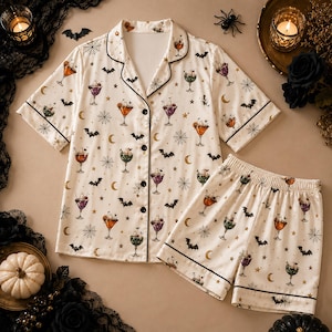 May include: A cream-colored short-sleeve pajama set featuring a repeating pattern of colorful cocktails, black bats, spiderwebs, stars, and crescent moons. The button-down shirt has a notched collar and black piping, while the shorts have an elastic waistband.