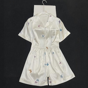May include: A white satin pajama set consisting of a short-sleeved button-down shirt with a collar and a chest pocket, paired with matching elastic-waist shorts. The fabric features a repeating pattern of small, colorful butterflies throughout.