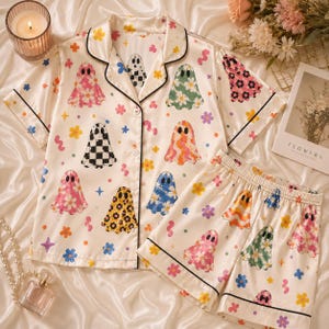 May include: A short-sleeve button-down pajama shirt and matching shorts set in cream fabric featuring a repeating pattern of colorful ghosts with various floral, checkered, and wavy designs, accented by small flowers and stars with black piping.