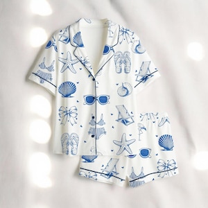 May include: White pajama set with a blue beach-themed pattern. The short-sleeved top and shorts feature illustrations of seashells, sunglasses, and beach chairs. The set has navy blue trim.
