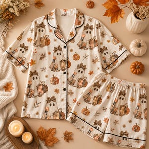 May include: A two-piece pajama set featuring a short-sleeve button-down shirt and matching shorts. The fabric is cream-colored with a repeating pattern of ghosts wearing leopard print bows, orange and brown plaid pumpkins, and autumn leaves.