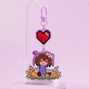 May include: A purple keychain featuring a cartoon character with brown hair and a flower crown, sitting in a bed of yellow flowers. Above the character is a red pixelated heart. The keychain is suspended from a purple clasp.