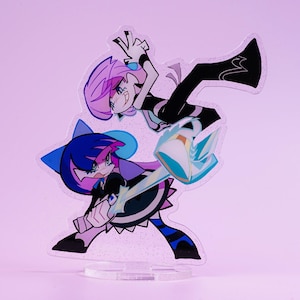 Könnte beinhalten: Ein klarer Acrylständer mit zwei Anime-Charakteren in dynamischen Posen. Die Charaktere haben rosa und blaue Haare und tragen schwarz-weiße Outfits mit blauen und violetten Akzenten. Der Ständer steht vor einem hellrosa Hintergrund.