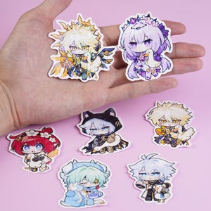 Könnte beinhalten: Eine Sammlung farbenfroher Aufkleber im Anime-Stil mit verschiedenen Charakteren. Die Aufkleber zeigen detaillierte Illustrationen mit leuchtenden Farben und einzigartigen Designs. Die Aufkleber werden in einer Hand vor einem rosa Hintergrund gehalten.