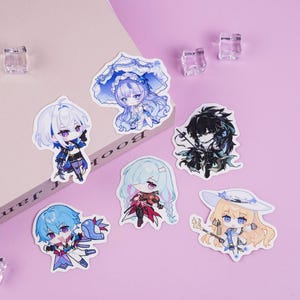 May include: A collection of anime-style character stickers with various designs. The stickers feature characters with blue, white, and black hair, wearing detailed outfits. The stickers are on a pink background with a book and clear decorative cubes.