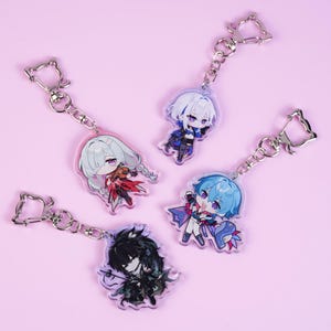 May include: Four anime-style character keychains with silver-colored hardware. Each keychain features a different character design with varying hair colors, outfits, and poses. The keychains are on a light pink background.