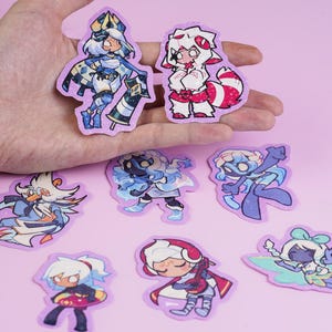 May include: A collection of colorful cartoon character stickers on a pink background. The stickers feature various anime-style figures in different poses and outfits, with a variety of colors including blue, red, white, and purple. The stickers are held in a hand.