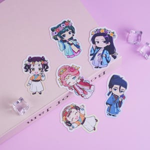 Può includere: Una collezione di sette adesivi in stile anime raffiguranti personaggi con fasce floreali e abiti tradizionali. Gli adesivi, in vari colori tra cui blu, rosa e arancione, sono disposti su una superficie rosa chiaro, con decorazioni quadrate trasparenti.