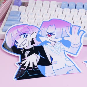 Puede incluir: Una pegatina con dos personajes de estilo anime con pelo rosa y azul, vestidos con trajes. Un personaje lleva un traje negro, el otro un traje blanco. La pegatina está sobre una superficie rosa cerca de un teclado.