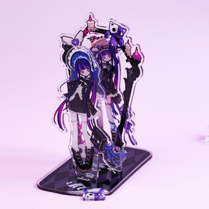 Puede incluir: Figura acrílica con dos personajes de estilo anime con pelo largo y colorido y atuendos de inspiración gótica. Las figuras están sobre una base a cuadros en blanco y negro, con elementos decorativos como velas y un gato morado. El diseño general tiene una estética oscura y caprichosa.