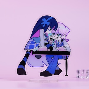 May include: Acrylic figure featuring two anime-style characters in blue and purple. One character has long dark blue hair and the other has light purple hair and glasses. They are positioned behind a black table, with a light pink background.