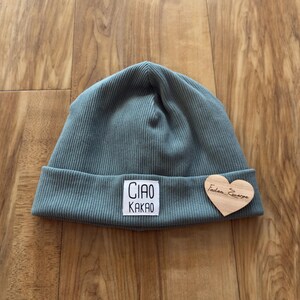 Könnte beinhalten: Eine tealfarbene Rippstrickmütze mit umgeschlagenem Saum. Ein weißes quadratisches Patch zeigt die Worte "CIAO KAKAO" in Schwarz. Ein herzförmiges Holzschild ist angebracht.