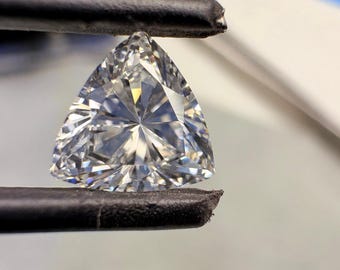 Può includere: Una gemma trasparente, tagliata a forma di triangolo, tenuta da pinzette nere. La gemma ha molte sfaccettature che riflettono la luce, creando un effetto scintillante. La forma è un perfetto triangolo equilatero, mostrando la sua chiarezza.
