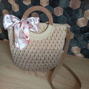 May include: A beige crocheted handbag with a wooden handle and a floral patterned silk scarf tied to it. The bag has a detachable, light brown leather strap and a gold-colored clasp. The text "Milla handmade" is visible at the bottom.