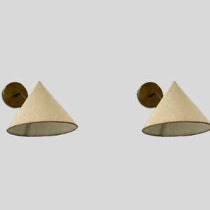 Peut inclure: Deux appliques murales avec des abat-jours coniques. Les abat-jours sont de couleur beige clair avec une bordure foncée. Les appliques ont une base circulaire de couleur laiton. Fond gris clair.