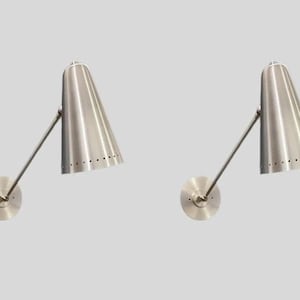 Op de afbeelding: Twee wandkandelaars van geborsteld nikkel. Elke wandkandelaar heeft een conische kap met een rij kleine gaatjes, een verstelbare arm en een ronde wandbevestiging. De lampen zijn ontworpen voor een moderne uitstraling.