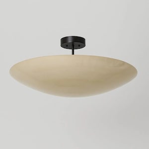 May include: A contemporary ceiling light with a cream-coloured, saucer-shaped shade. The shade is attached to a black base and stem. The light fixture is designed for indoor use and provides ambient lighting.