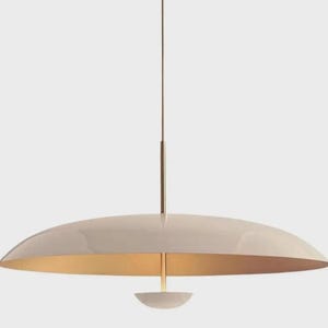May include: A modern, cream-colored pendant light fixture with a wide, saucer-shaped shade. The interior of the shade is gold, and a small, white, bowl-shaped element hangs below. The fixture is suspended by a thin, bronze-colored rod.