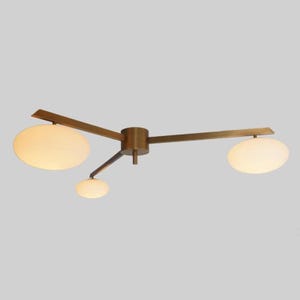 May include: A modern ceiling light fixture with a brass finish. The light has three arms, each with an oval-shaped, frosted glass shade. The fixture is designed to be mounted on the ceiling and provides ambient lighting.
