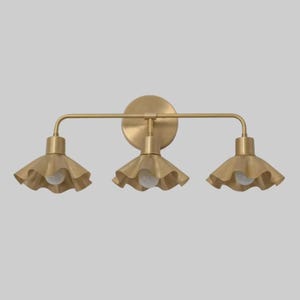 May include: A three-light vanity fixture with a brass finish. The fixture has a circular backplate, a horizontal bar, and three bell-shaped shades with ruffled edges. Each shade houses a single light bulb.