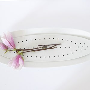 Op de afbeelding: Een witte ovale keramische schaal met een geperforeerde inzet. De schaal heeft een verhoogde rand en is versierd met een rij kleine gaatjes rond de rand. Een tak met roze magnolia bloemen ligt in de schaal.