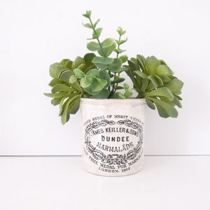 May include: White ceramic jar with black lettering that reads "Grand Medal of Merit Vienna. James Keiller & Sons. Dundee Marmalade. First Prize Medal for Marmalade. London. 1862". The jar is filled with green succulents.