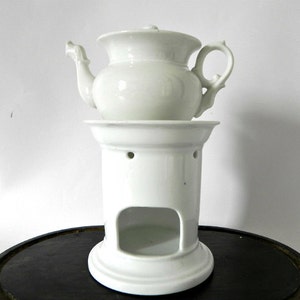 May include: White porcelain teapot with a lid and a handle, sitting on a white porcelain stand with holes. The stand has a round base and a hollow center.