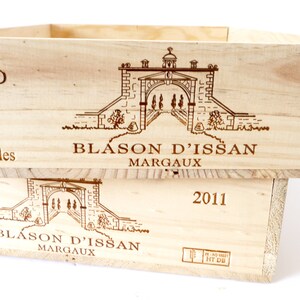 Op de afbeelding: Een houten wijnkist met de woorden "Blason d'Issan Margaux" en het jaar "2011" erop gedrukt. De kist heeft een tekening van een poort met een toren erop.