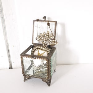 Op de afbeelding: Een klein, antiek glazen juwelendoosje met een scharnierend deksel en sierlijke metalen accenten. Het doosje is open en laat een delicate, met kristallen bezette broche zien.